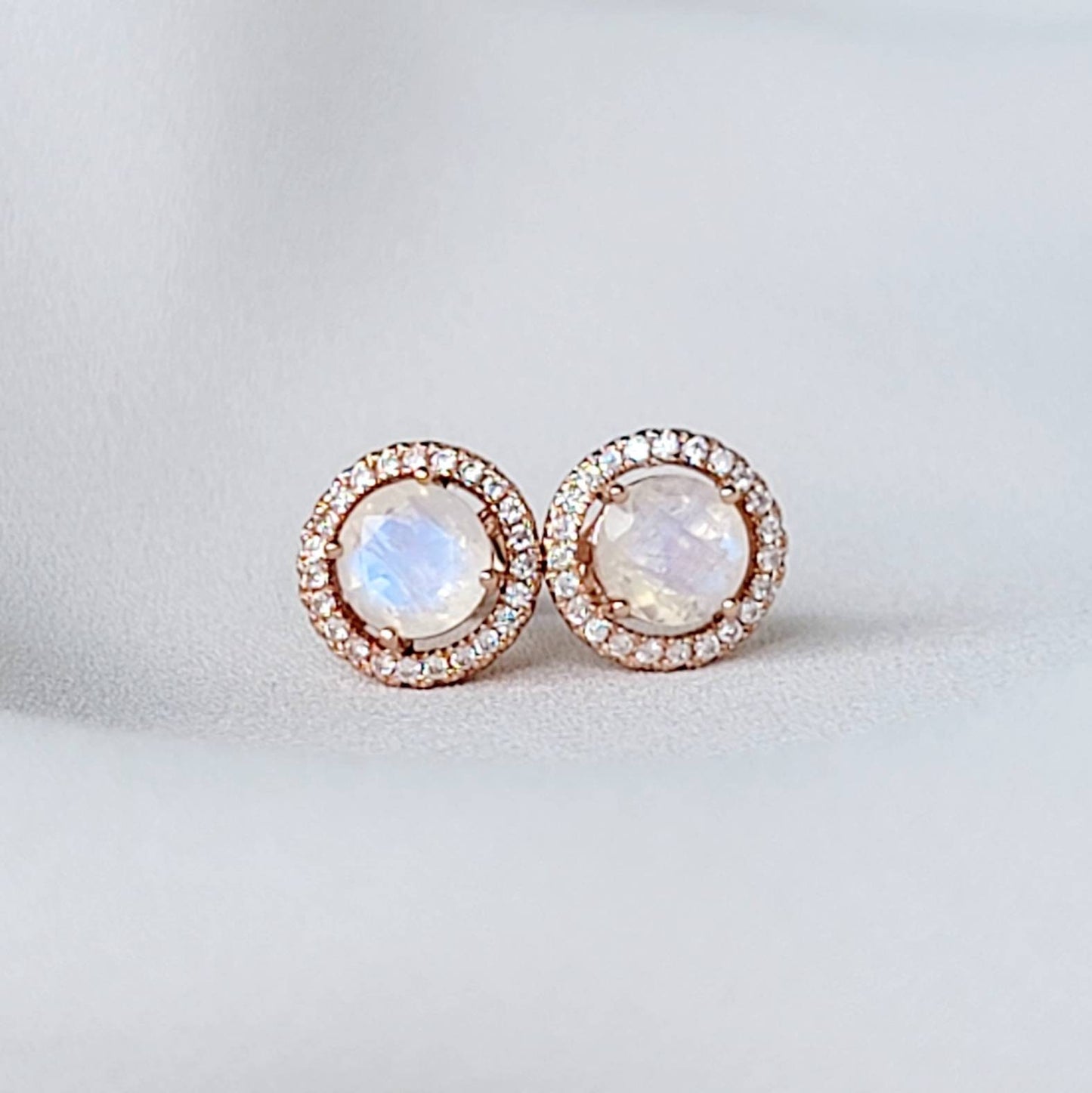 A pair of round, rose gold earrings with a rainbow moonstone center stone and a halo of smaller white stones