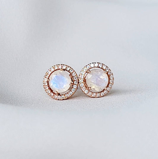 A pair of round, rose gold earrings with a rainbow moonstone center stone and a halo of smaller white stones