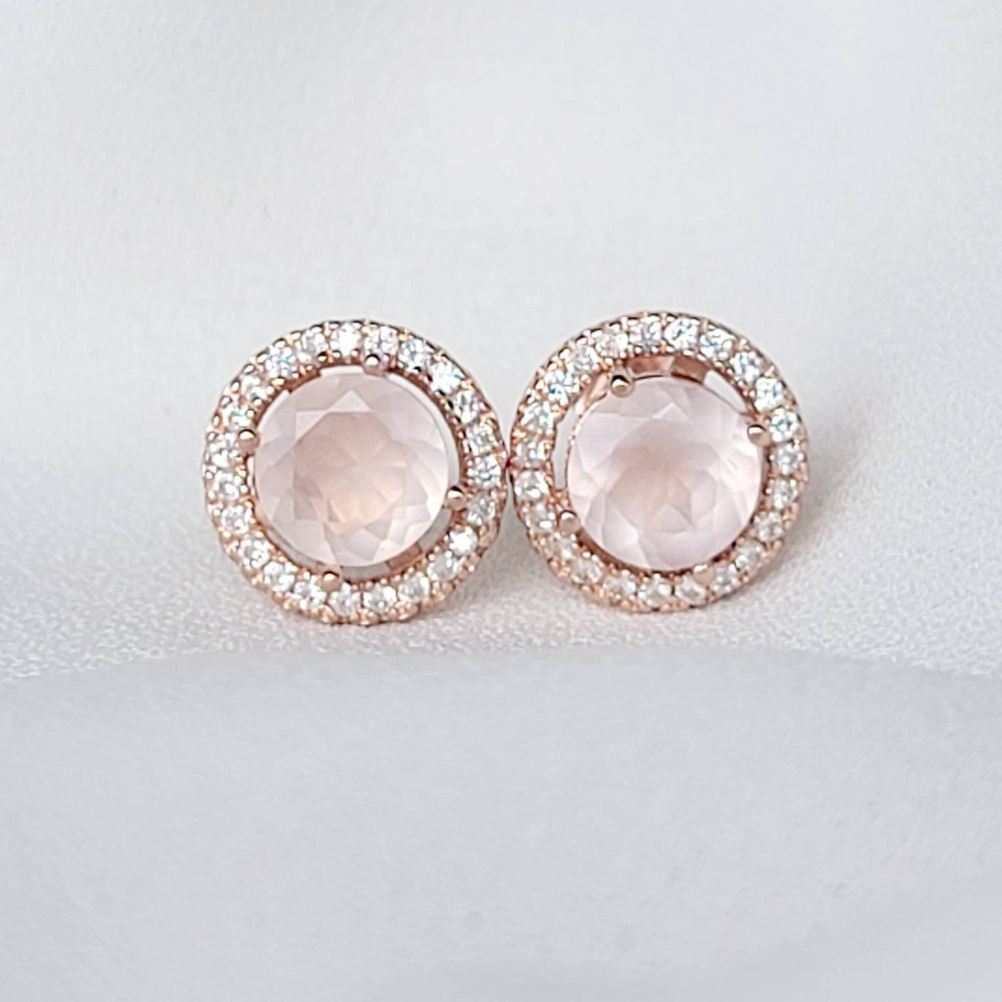 A pair of rose gold earrings with a round, light pink rose quartz gemstone surrounded by a halo of small, clear gemstones.