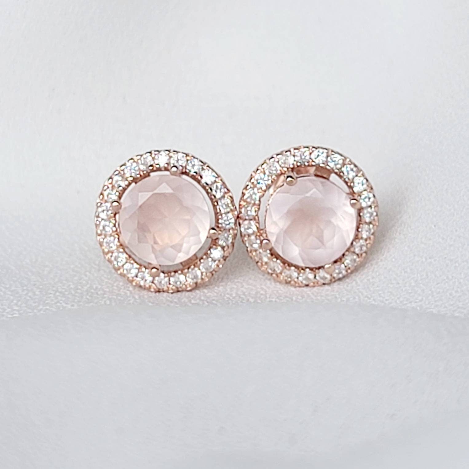 A pair of rose gold earrings with a round, light pink rose quartz gemstone surrounded by a halo of small, clear gemstones.