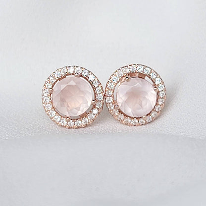 A pair of rose gold earrings with a round, light pink rose quartz gemstone surrounded by a halo of small, clear gemstones.