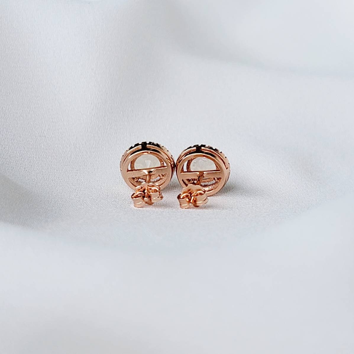 A pair of rose gold earrings with a round, light pink rose quartz gemstone surrounded by a halo of small, clear gemstones.
