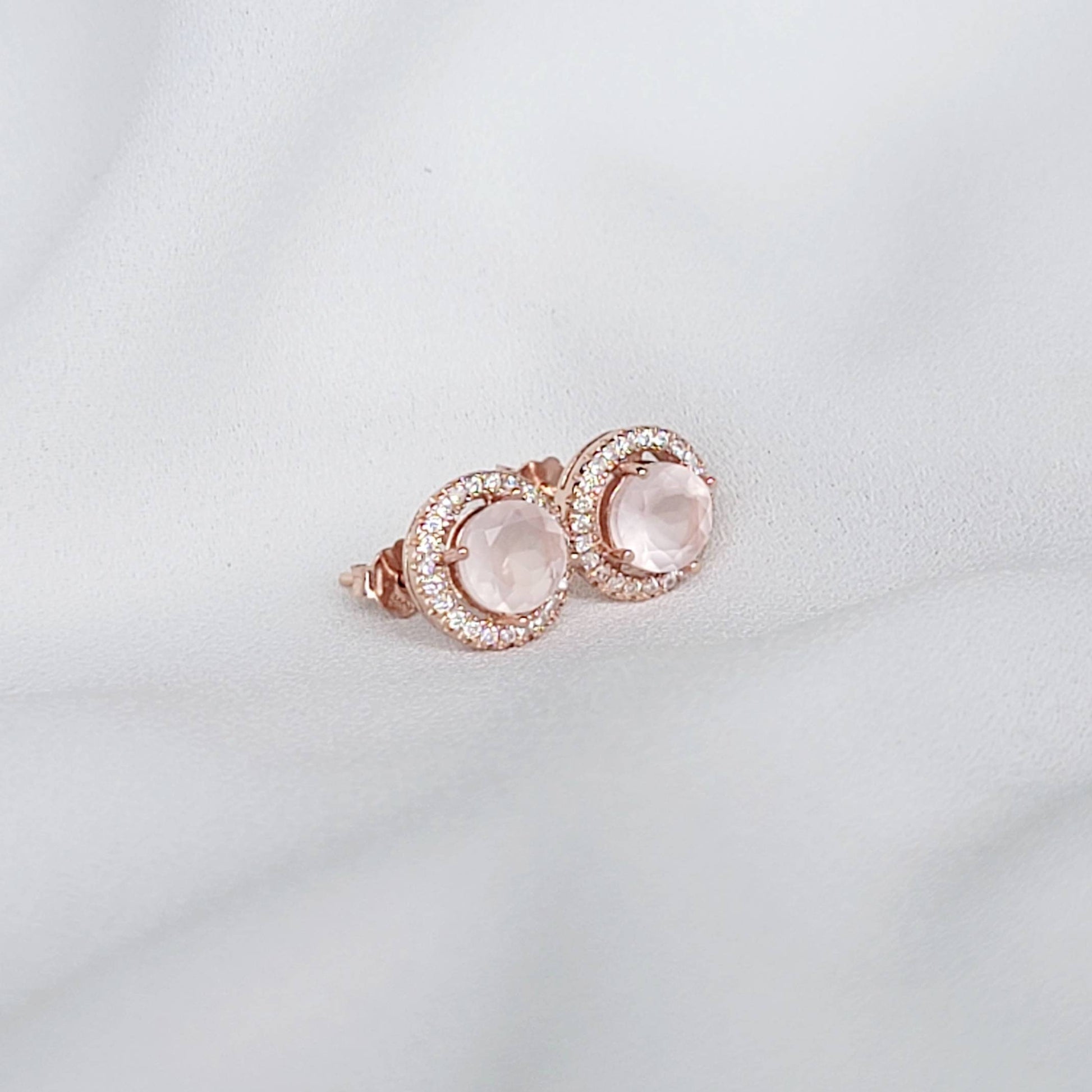 A pair of rose gold earrings with a round, light pink rose quartz gemstone surrounded by a halo of small, clear gemstones.