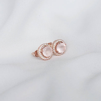 A pair of rose gold earrings with a round, light pink rose quartz gemstone surrounded by a halo of small, clear gemstones.