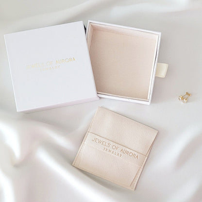 A white box with the text &#39;Jewels of Aurora Jewelry&#39; printed in gold. The box is open and a small, cream suede pouch with the same text printed in gold is inside. The pouch is sitting on a white surface.