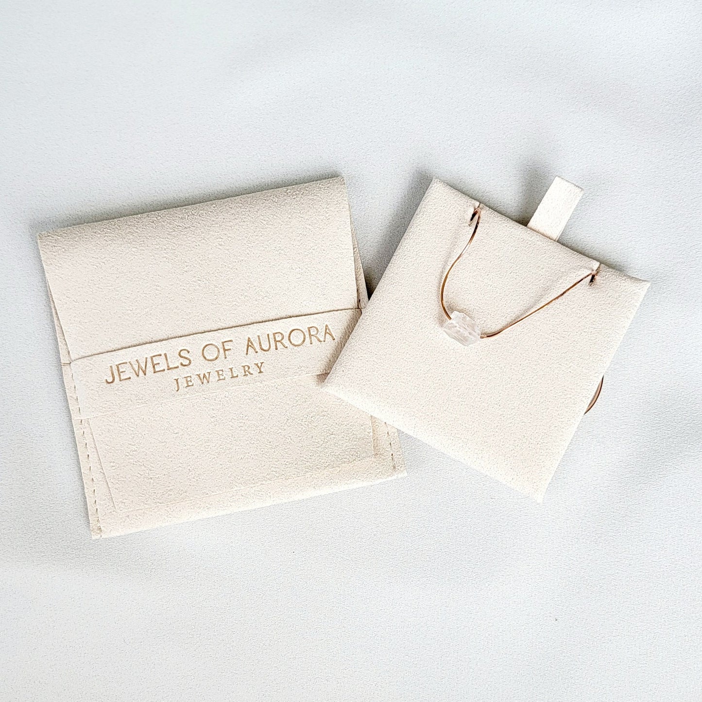 One cream velvet jewelry pouche. The larger pouch has the text &#39;Jewels of Aurora Jewelry&#39; printed on it. The smaller pouch contains a gold chain with a white crystal pendant.