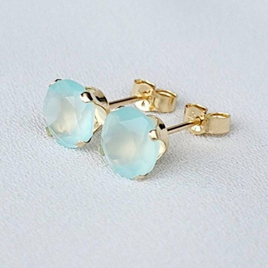 Pair of gold stud earrings featuring light blue, faceted gemstone accents.  Each earring showcases a round aqua stone set in a four-prong gold setting. The posts and butterfly clasps are also gold. These minimalist earrings are perfect for everyday wear or special occasions.