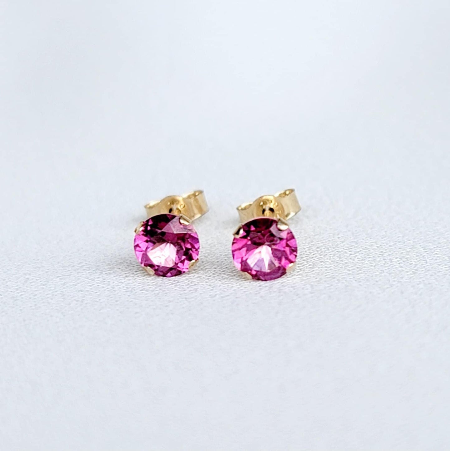 Pair of gold stud earrings featuring round, fuchsia-colored gemstones known at pink topaz.  The earrings have a simple, classic design, suitable for everyday wear or special occasions. The vibrant pink stones are securely set in gold posts, offering a touch of elegance and sophistication. These small, delicate earrings are perfect for adding a pop of color to any outfit.
