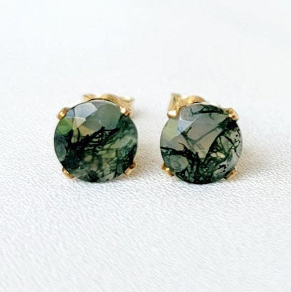 Pair of gold stud earrings featuring moss agate, faceted gemstone accents.  Each earring showcases a round clear agate stones with moss growing through it. It is set in a four-prong gold setting. The posts and butterfly clasps are also gold. These minimalist earrings are perfect for everyday wear or special occasions.