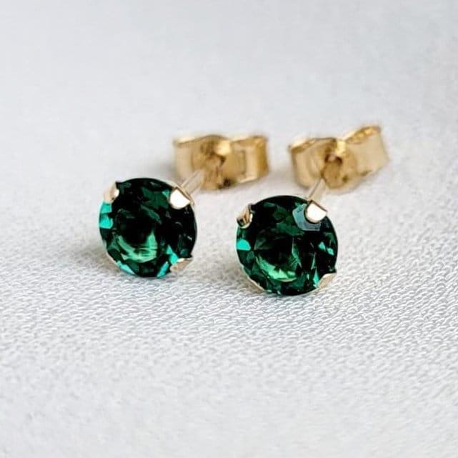 Close-up of a pair of gold stud earrings featuring sustainable emerald gemstones. The earrings have a classic, simple design with a four-prong setting for each stone. The gemstones appear to be emerald green, exhibiting brilliance and clarity. The gold metal appears smooth and polished. The earrings are presented against a neutral, light background.