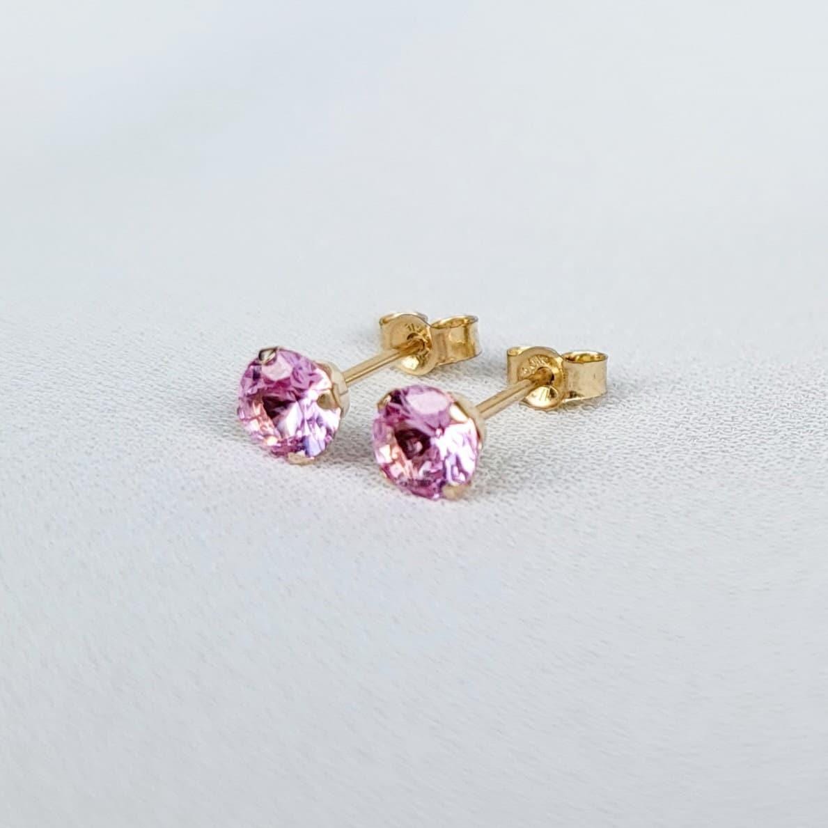 Close-up view of a pair of gold stud earrings. Each earring features a small, round, light pink sapphire gemstones. The earrings have a simple, classic design, suitable for everyday wear. The gemstones appear to be sparkling and have a smooth surface. The gold settings are delicate and complement the pink stones. The earrings are displayed on a white textured surface.