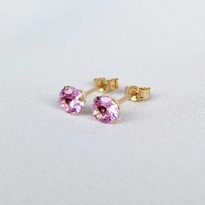 Close-up view of a pair of gold stud earrings. Each earring features a small, round, light pink sapphire gemstones. The earrings have a simple, classic design, suitable for everyday wear. The gemstones appear to be sparkling and have a smooth surface. The gold settings are delicate and complement the pink stones. The earrings are displayed on a white textured surface.