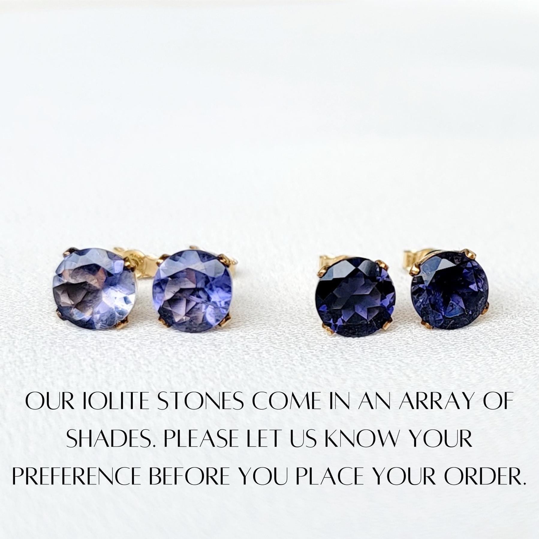 Four round iolite gemstone earrings with gold settings. The earrings are displayed on a white surface. The text below the earrings reads: &#39;OUR IOLITE STONES COME IN AN ARRAY OF SHADES. PLEASE LET US KNOW YOUR PREFERENCE BEFORE YOU PLACE YOUR ORDER.&#39;
