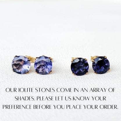 Four round iolite gemstone earrings with gold settings. The earrings are displayed on a white surface. The text below the earrings reads: &#39;OUR IOLITE STONES COME IN AN ARRAY OF SHADES. PLEASE LET US KNOW YOUR PREFERENCE BEFORE YOU PLACE YOUR ORDER.&#39;