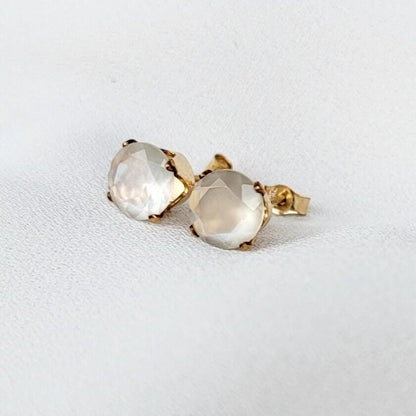 Close-up of a pair of gold stud earrings featuring round, faceted, shimmery gray moonstone gemstones. The earrings have a simple, elegant design, with the gemstones securely set in gold prongs. The gemstones have a subtle shimmer and appear to be translucent. These minimalist earrings would be a delicate addition to any jewelry collection.