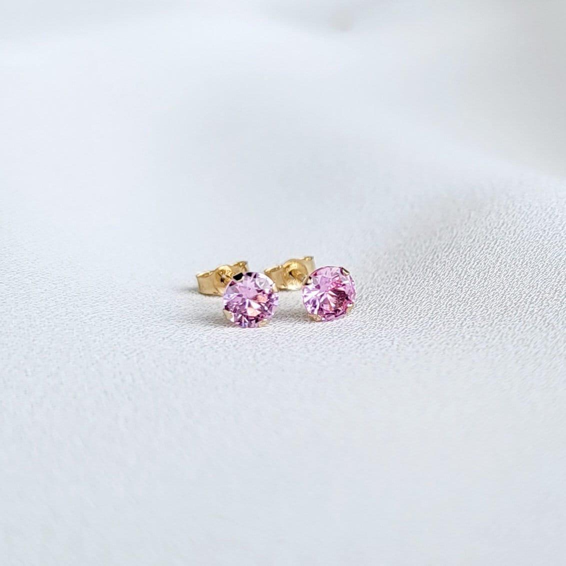 Close-up view of a pair of gold stud earrings. Each earring features a small, round, light pink sapphire gemstones. The earrings have a simple, classic design, suitable for everyday wear. The gemstones appear to be sparkling and have a smooth surface. The gold settings are delicate and complement the pink stones. The earrings are displayed on a white textured surface.