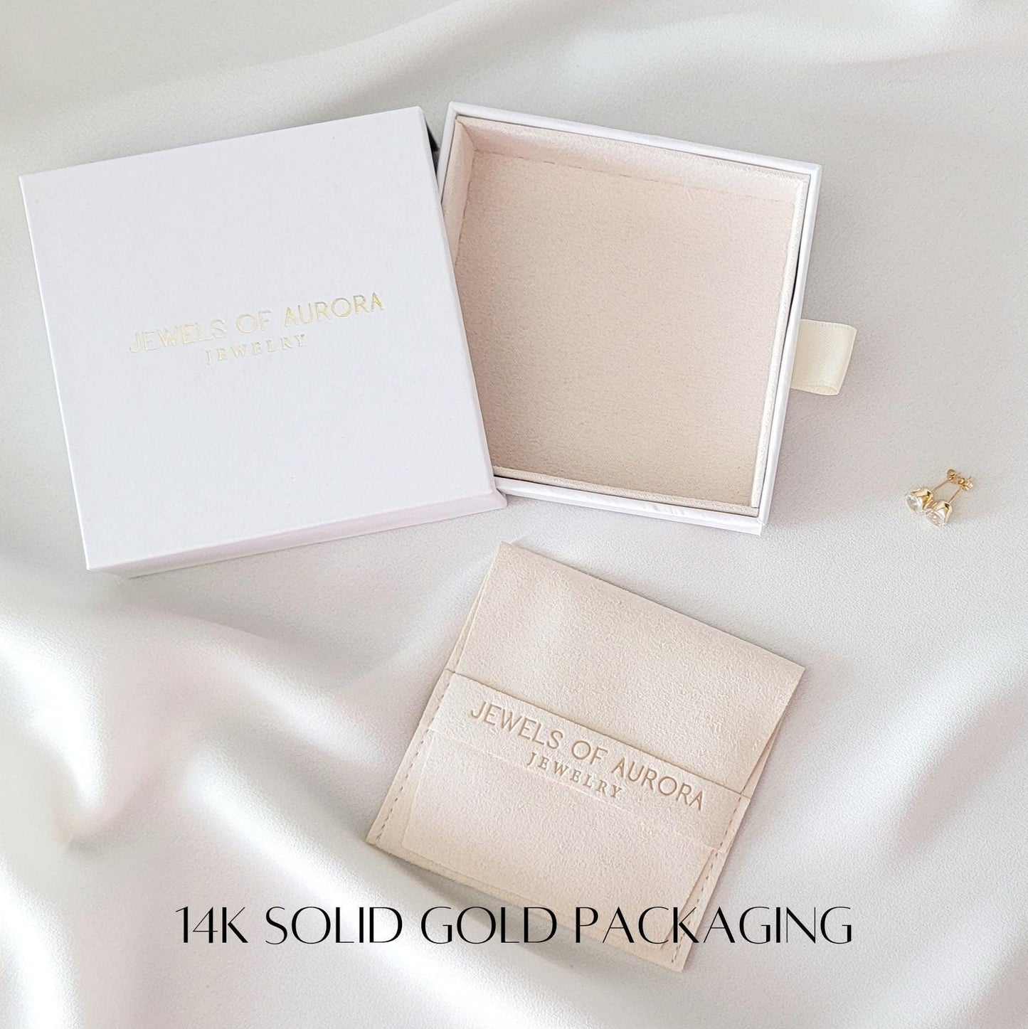14k solid gold packaging.  White jewelry boxes with the logo &#39;Jewels of Aurora Jewelry&#39; are shown.  A smaller, off-white suede pouch with the same logo is also included.  A pair of gold earrings is displayed next to the boxes.
