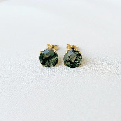 Pair of gold stud earrings featuring moss agate, faceted gemstone accents.  Each earring showcases a round clear agate stones with moss growing through it. It is set in a four-prong gold setting. The posts and butterfly clasps are also gold. These minimalist earrings are perfect for everyday wear or special occasions.