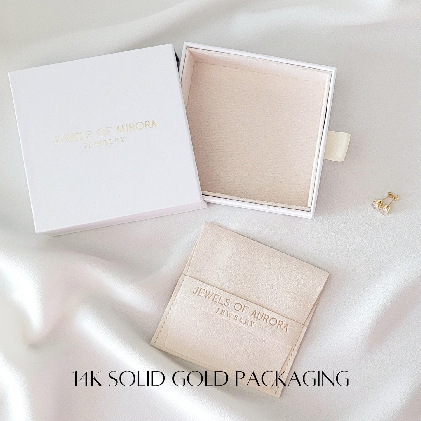 A white box with the text &#39;Jewels of Aurora Jewelry&#39; on the top. The box is open and the inside is lined with white velvet. A smaller white box with a beige velvet lining is inside the larger box. A small beige velvet pouch with the text &#39;Jewels of Aurora Jewelry&#39; is on a white surface. The text &#39;14k Solid Gold Packaging&#39; is printed on the surface.