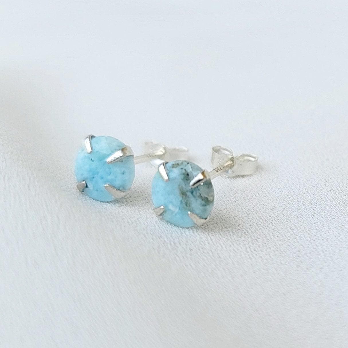 A pair of silver stud earrings with light blue Larimar round gemstones. The gemstones have a speckled pattern.
