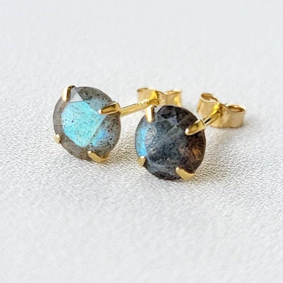 A pair of gold stud earrings with round, iridescent labradorite gemstones. The gemstones have a dark gray base with blue and green flashes of color.
