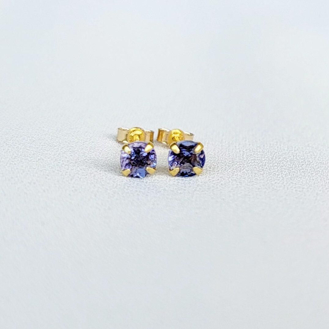 Close-up view of a pair of gold tanzanite stud earrings, each featuring a small, round, purple gemstone. The earrings have a simple, classic design, with the gemstones securely set in the gold settings. The gemstones appear to be a light to medium shade of purple. The gold is a warm, yellowish tone. The earrings are displayed against a plain, off-white background.
