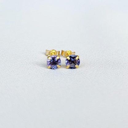 Close-up view of a pair of gold tanzanite stud earrings, each featuring a small, round, purple gemstone. The earrings have a simple, classic design, with the gemstones securely set in the gold settings. The gemstones appear to be a light to medium shade of purple. The gold is a warm, yellowish tone. The earrings are displayed against a plain, off-white background.