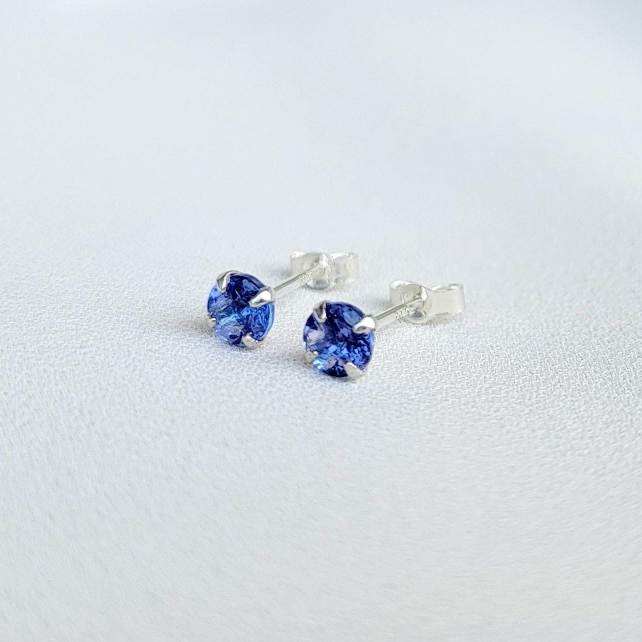 Close-up view of a pair of gold tanzanite stud earrings, each featuring a small, round, purple gemstone. The earrings have a simple, classic design, with the gemstones securely set in the gold settings. The gemstones appear to be a light to medium shade of purple. The gold is a warm, yellowish tone. The earrings are displayed against a plain, off-white background.