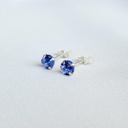 Close-up view of a pair of gold tanzanite stud earrings, each featuring a small, round, purple gemstone. The earrings have a simple, classic design, with the gemstones securely set in the gold settings. The gemstones appear to be a light to medium shade of purple. The gold is a warm, yellowish tone. The earrings are displayed against a plain, off-white background.