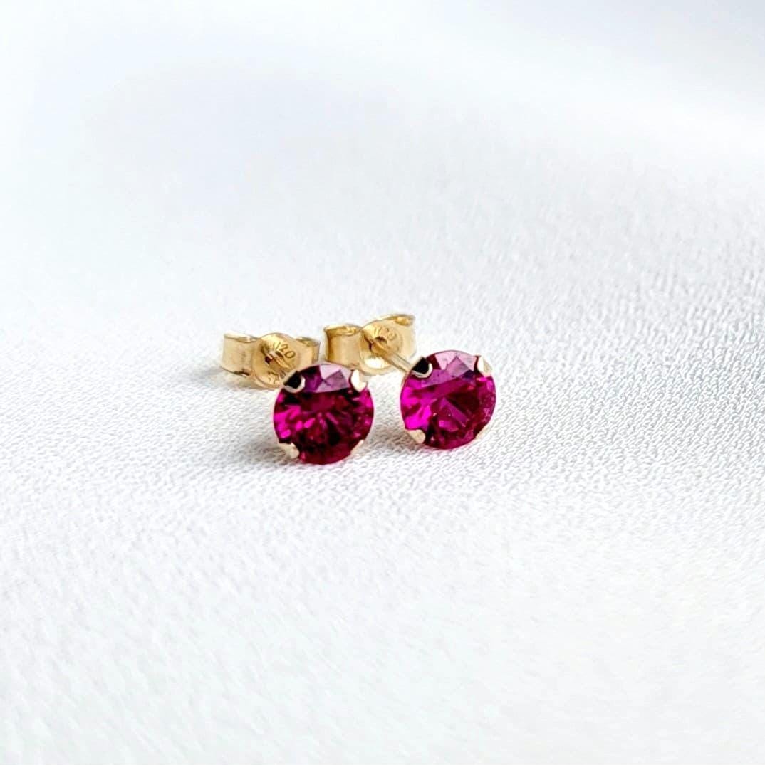 A pair of gold stud earrings with fascia colored ruby gemstones. The earrings are small and round, with a simple design.