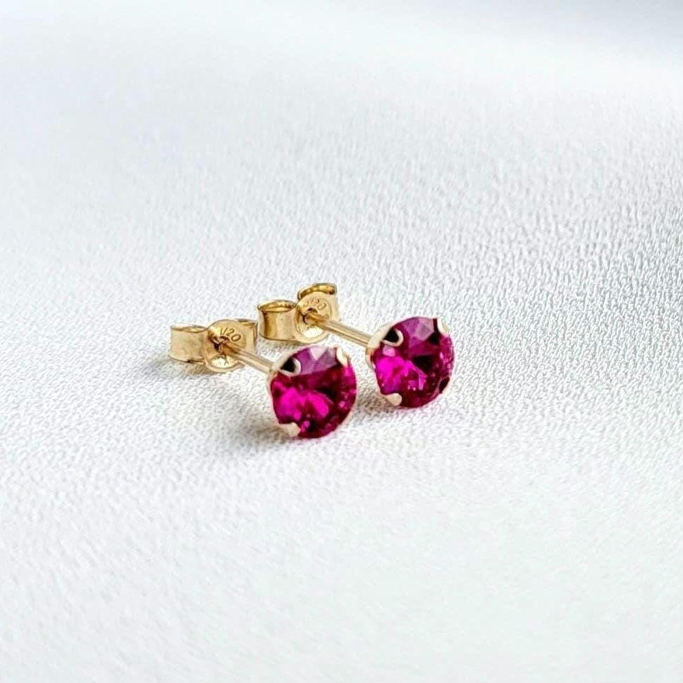 A pair of gold stud earrings with fascia colored ruby gemstones. The earrings are small and round, with a simple design.