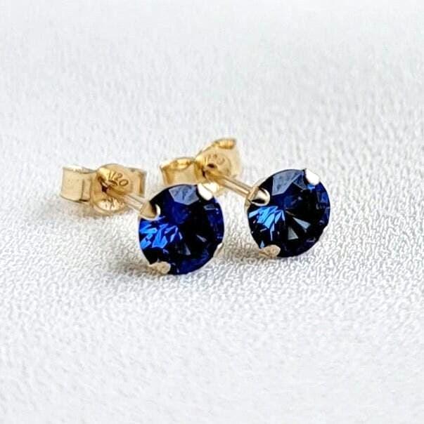 A pair of gold stud earrings with round faceted blue sapphire gemstones. The earrings are on a white surface.