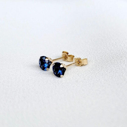 A pair of gold stud earrings with round faceted blue sapphire gemstones. The earrings are on a white surface.