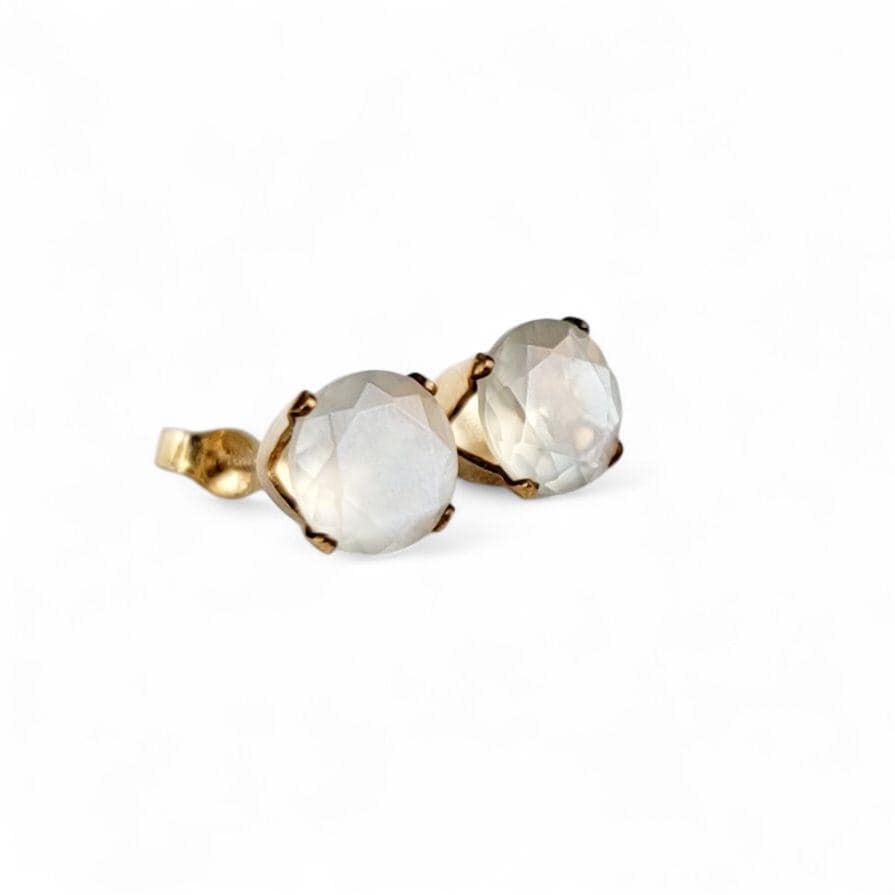 Close-up of a pair of gold stud earrings featuring round, faceted, shimmery gray moonstone gemstones. The earrings have a simple, elegant design, with the gemstones securely set in gold prongs. The gemstones have a subtle shimmer and appear to be translucent. These minimalist earrings would be a delicate addition to any jewelry collection.