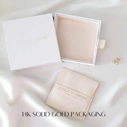 14k solid gold packaging.  White jewelry boxes with the logo &#39;Jewels of Aurora Jewelry&#39; are shown.  A beige suede pouch with the same logo is also included.  A pair of gold earrings is displayed next to the boxes.