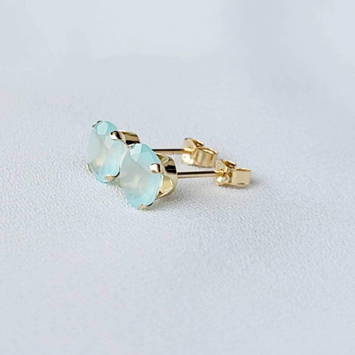 Pair of gold stud earrings featuring light blue, faceted gemstone accents.  Each earring showcases a round aqua stone set in a four-prong gold setting. The posts and butterfly clasps are also gold. These minimalist earrings are perfect for everyday wear or special occasions.