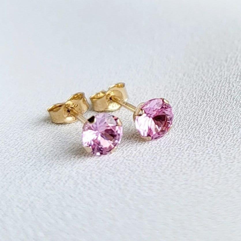 Close-up view of a pair of gold stud earrings. Each earring features a small, round, light pink sapphire gemstones. The earrings have a simple, classic design, suitable for everyday wear. The gemstones appear to be sparkling and have a smooth surface. The gold settings are delicate and complement the pink stones. The earrings are displayed on a white textured surface.
