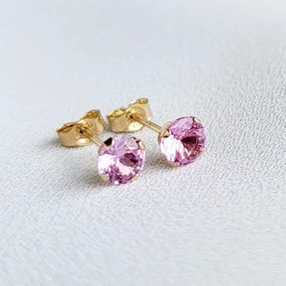 Close-up view of a pair of gold stud earrings. Each earring features a small, round, light pink sapphire gemstones. The earrings have a simple, classic design, suitable for everyday wear. The gemstones appear to be sparkling and have a smooth surface. The gold settings are delicate and complement the pink stones. The earrings are displayed on a white textured surface.