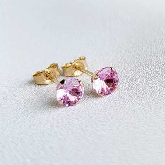 Close-up view of a pair of gold stud earrings. Each earring features a small, round, light pink sapphire gemstones. The earrings have a simple, classic design, suitable for everyday wear. The gemstones appear to be sparkling and have a smooth surface. The gold settings are delicate and complement the pink stones. The earrings are displayed on a white textured surface.