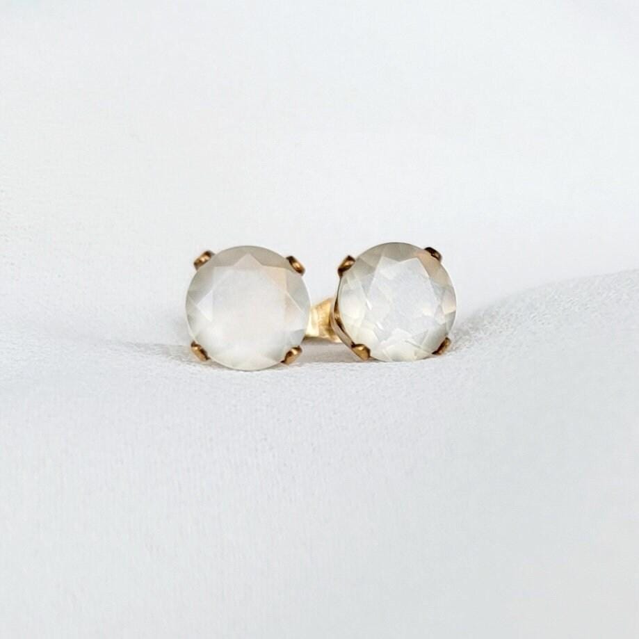 Close-up of a pair of gold stud earrings featuring round, faceted, shimmery gray moonstone gemstones. The earrings have a simple, elegant design, with the gemstones securely set in gold prongs. The gemstones have a subtle shimmer and appear to be translucent. These minimalist earrings would be a delicate addition to any jewelry collection.