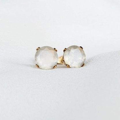 Close-up of a pair of gold stud earrings featuring round, faceted, shimmery gray moonstone gemstones. The earrings have a simple, elegant design, with the gemstones securely set in gold prongs. The gemstones have a subtle shimmer and appear to be translucent. These minimalist earrings would be a delicate addition to any jewelry collection.