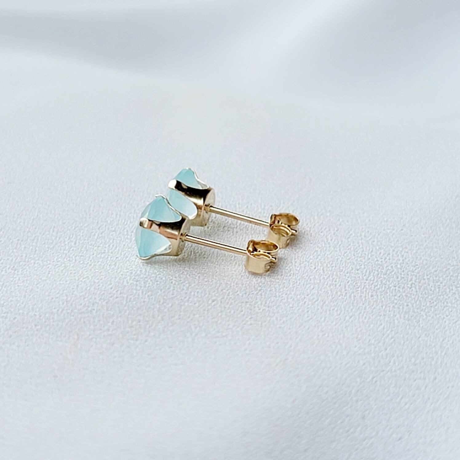 Pair of gold stud earrings featuring light blue, faceted gemstone accents.  Each earring showcases a round aqua stone set in a four-prong gold setting. The posts and butterfly clasps are also gold. These minimalist earrings are perfect for everyday wear or special occasions.