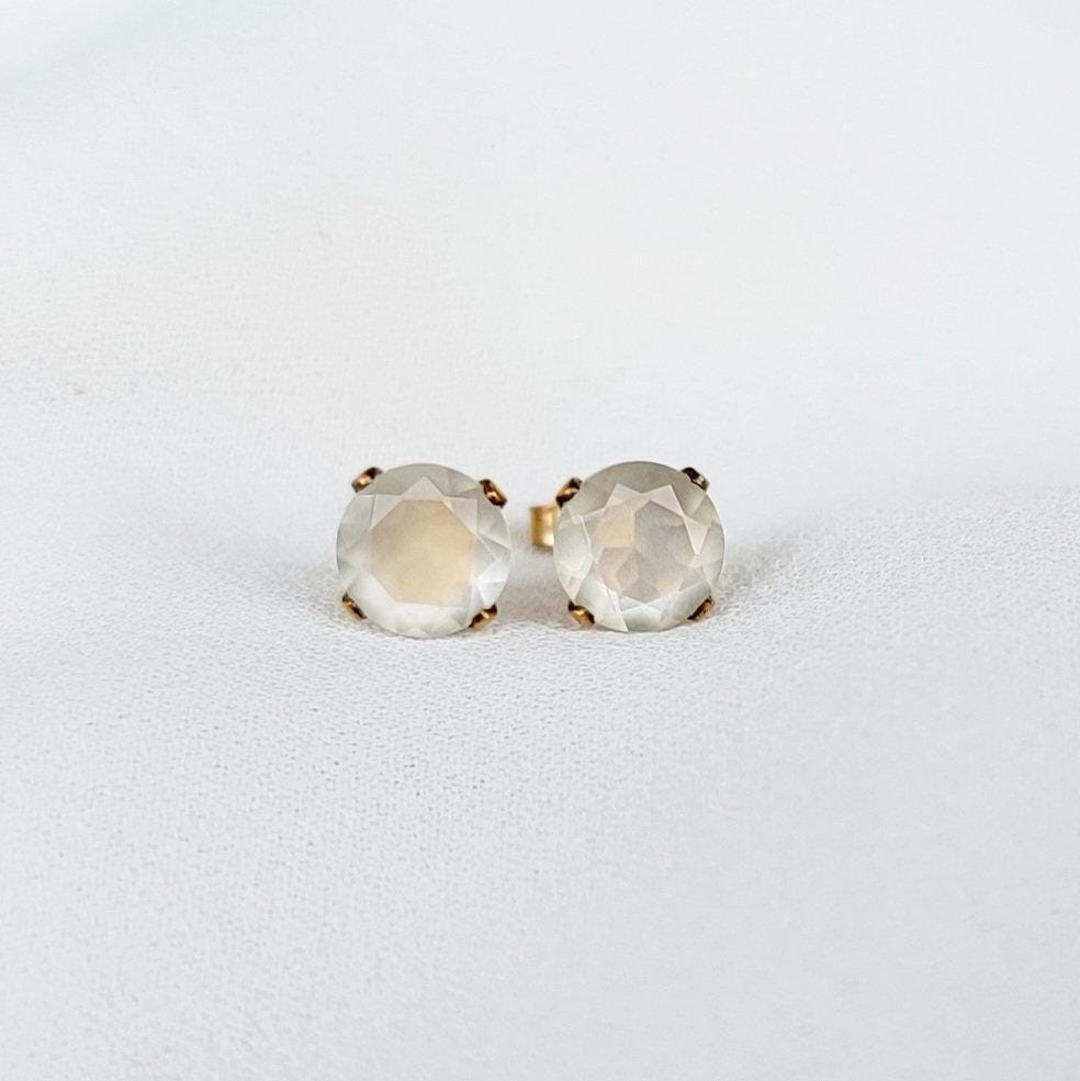 Close-up of a pair of gold stud earrings featuring round, faceted, shimmery gray moonstone gemstones. The earrings have a simple, elegant design, with the gemstones securely set in gold prongs. The gemstones have a subtle shimmer and appear to be translucent. These minimalist earrings would be a delicate addition to any jewelry collection.