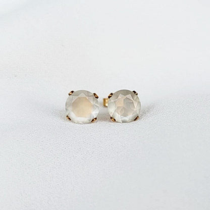 Close-up of a pair of gold stud earrings featuring round, faceted, shimmery gray moonstone gemstones. The earrings have a simple, elegant design, with the gemstones securely set in gold prongs. The gemstones have a subtle shimmer and appear to be translucent. These minimalist earrings would be a delicate addition to any jewelry collection.