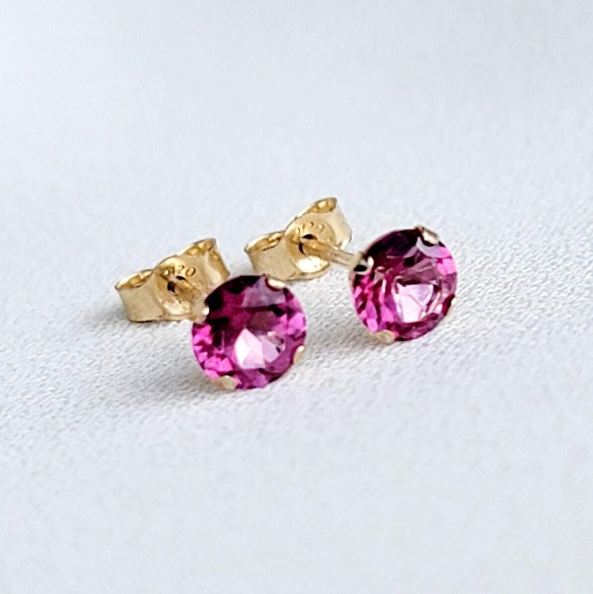 Pair of gold stud earrings featuring round, fuchsia-colored gemstones known at pink topaz.  The earrings have a simple, classic design, suitable for everyday wear or special occasions. The vibrant pink stones are securely set in gold posts, offering a touch of elegance and sophistication. These small, delicate earrings are perfect for adding a pop of color to any outfit.