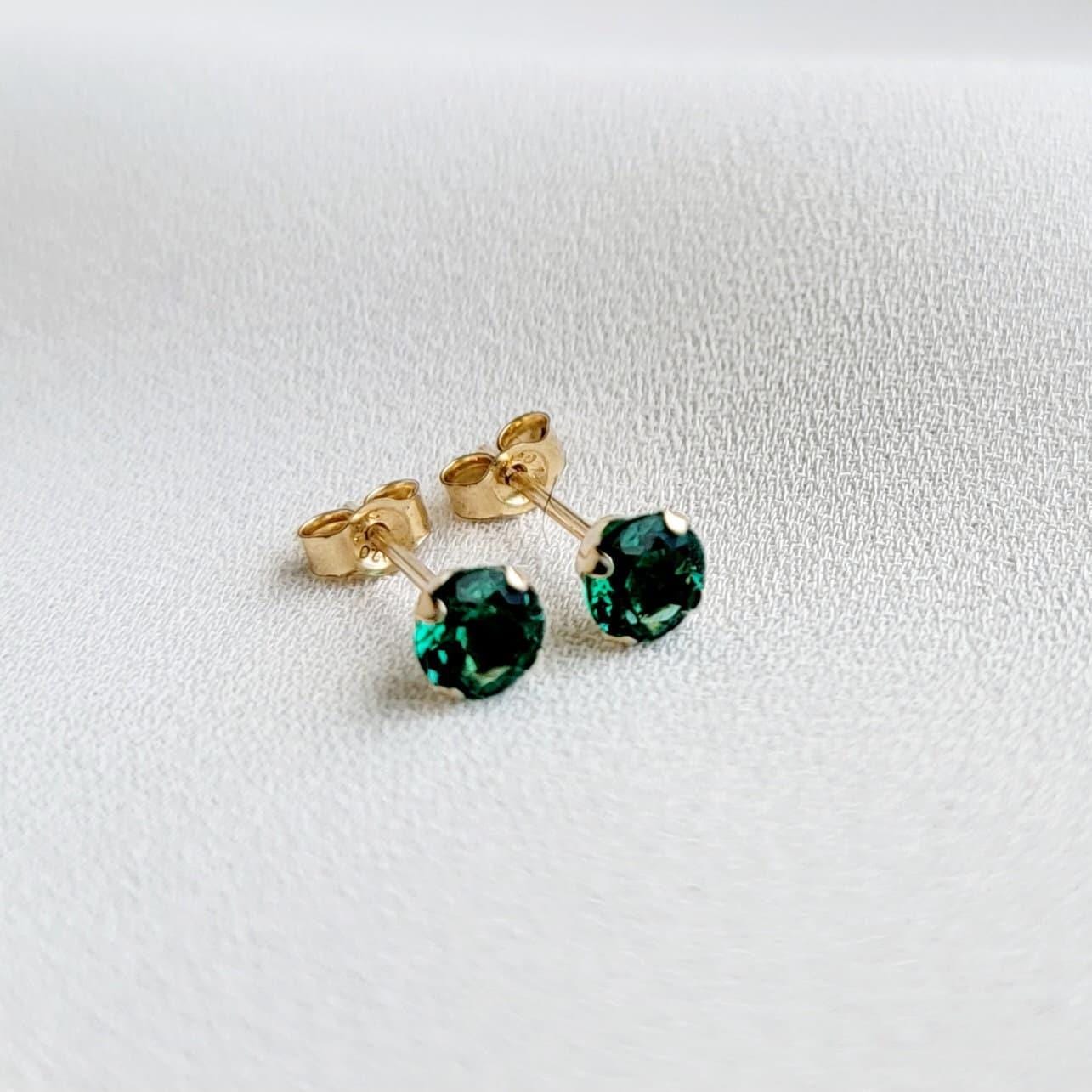 Close-up of a pair of gold stud earrings featuring sustainable emerald gemstones. The earrings have a classic, simple design with a four-prong setting for each stone. The gemstones appear to be emerald green, exhibiting brilliance and clarity. The gold metal appears smooth and polished. The earrings are presented against a neutral, light background.