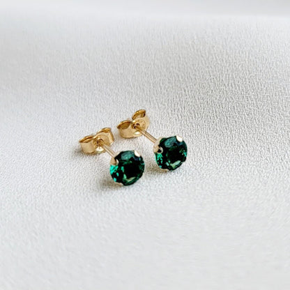 Close-up of a pair of gold stud earrings featuring sustainable emerald gemstones. The earrings have a classic, simple design with a four-prong setting for each stone. The gemstones appear to be emerald green, exhibiting brilliance and clarity. The gold metal appears smooth and polished. The earrings are presented against a neutral, light background.