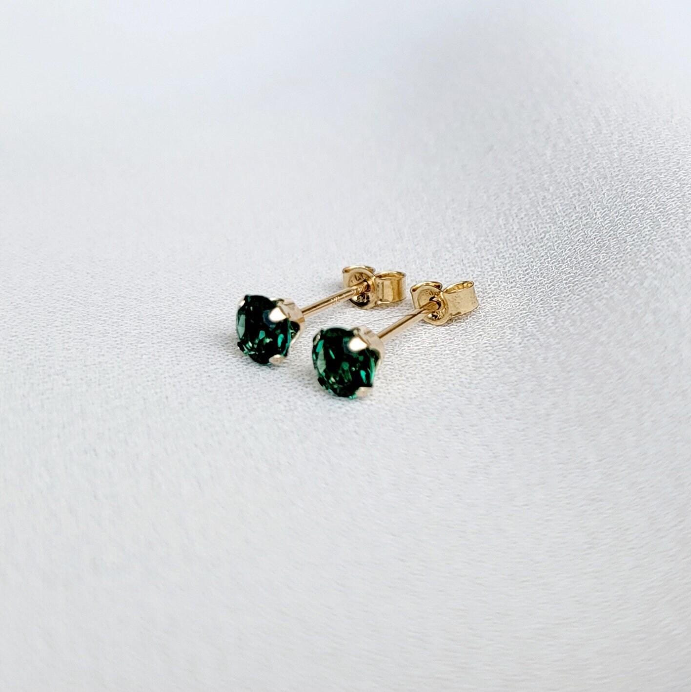 Close-up of a pair of gold stud earrings featuring sustainable emerald gemstones. The earrings have a classic, simple design with a four-prong setting for each stone. The gemstones appear to be emerald green, exhibiting brilliance and clarity. The gold metal appears smooth and polished. The earrings are presented against a neutral, light background.