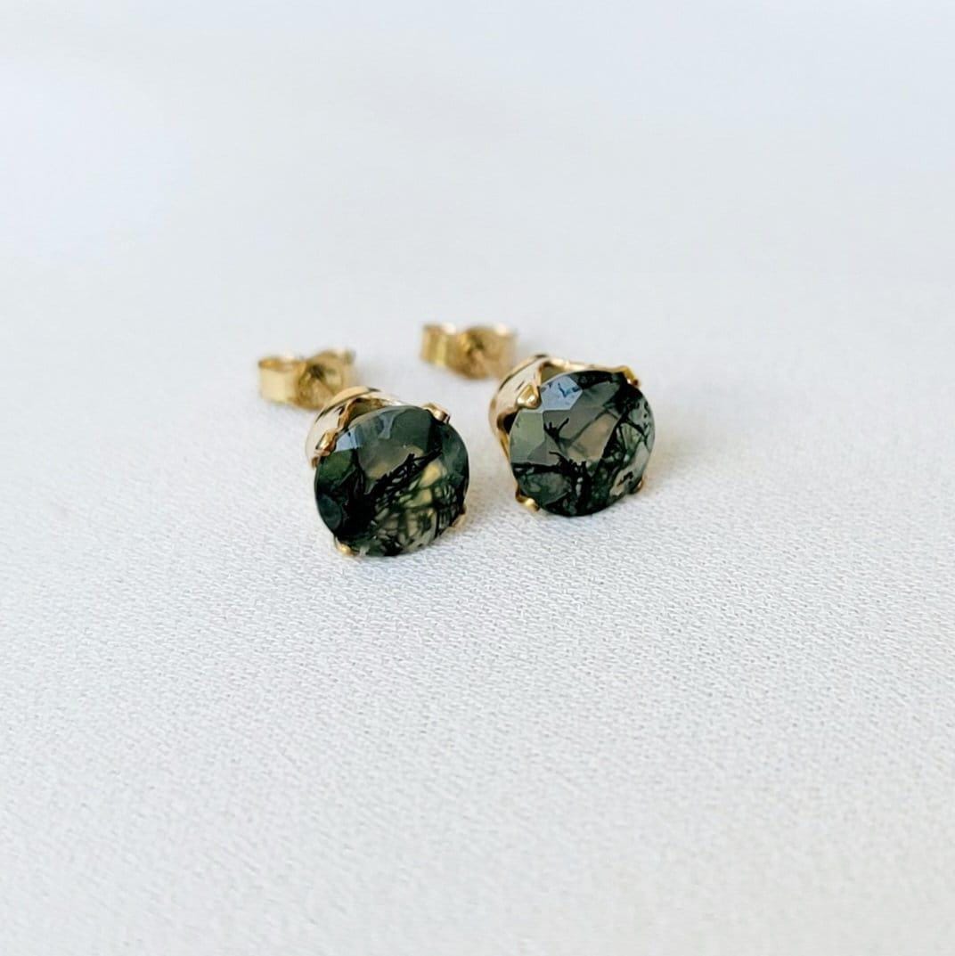 Pair of gold stud earrings featuring moss agate, faceted gemstone accents.  Each earring showcases a round clear agate stones with moss growing through it. It is set in a four-prong gold setting. The posts and butterfly clasps are also gold. These minimalist earrings are perfect for everyday wear or special occasions.