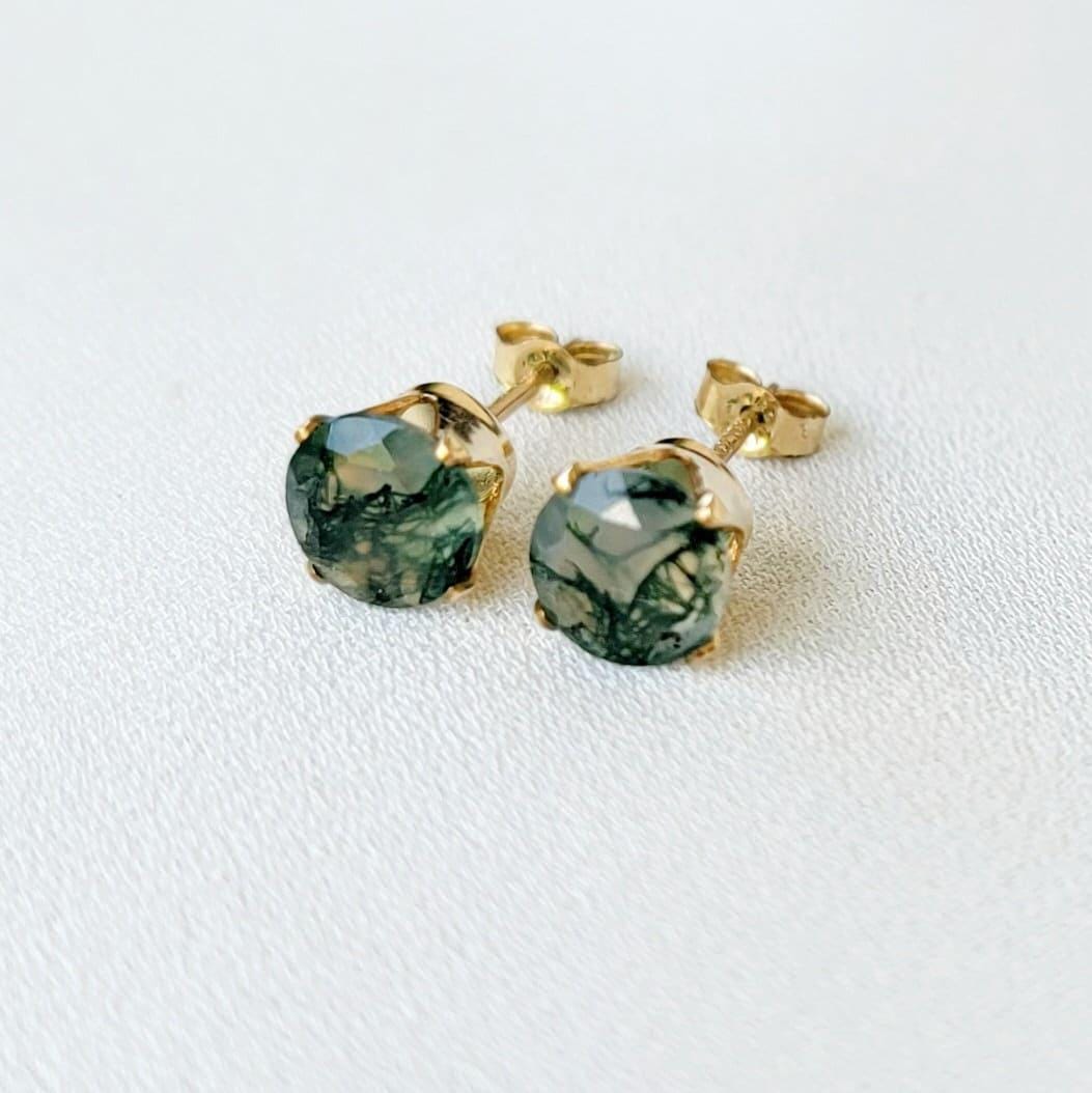 Pair of gold stud earrings featuring moss agate, faceted gemstone accents.  Each earring showcases a round clear agate stones with moss growing through it. It is set in a four-prong gold setting. The posts and butterfly clasps are also gold. These minimalist earrings are perfect for everyday wear or special occasions.